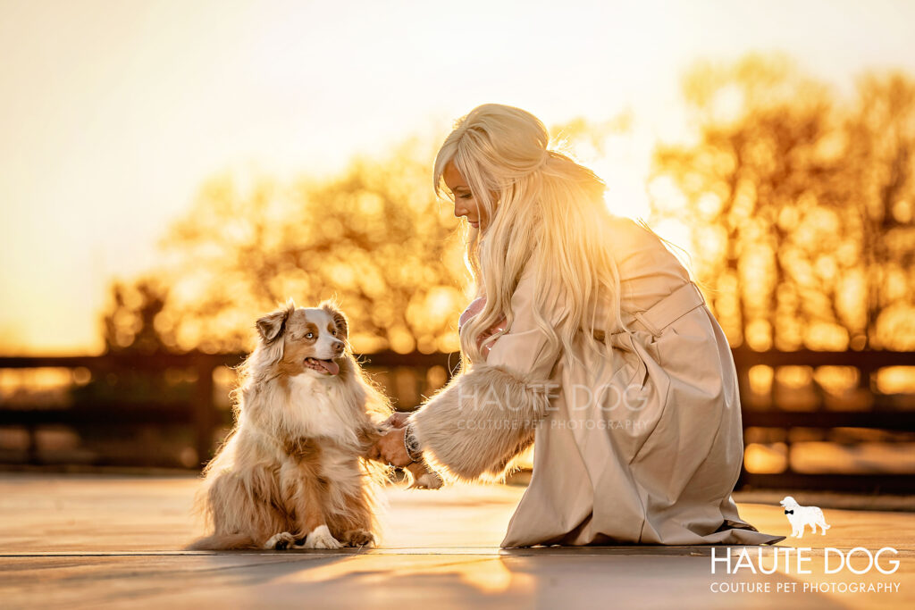 Blonde woman kneeling in golden sunlight holding her Australian Shepherd dog's paw.