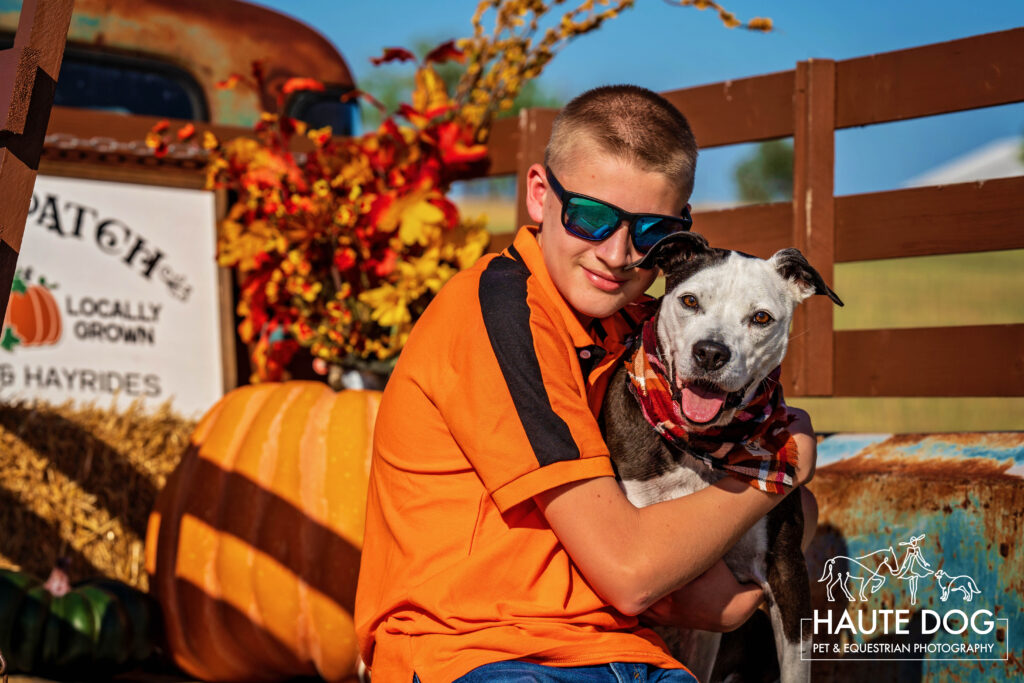A teenage boy hugs his black and white dog surrounded by pumpkins and fall decor.