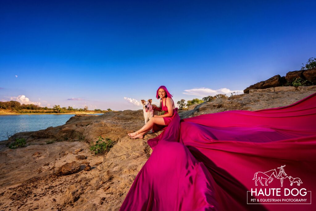 Woman with pink hair sits on the bluffs of Grapevine Lake with her dog while her pink flying dress flutters over the rocks.