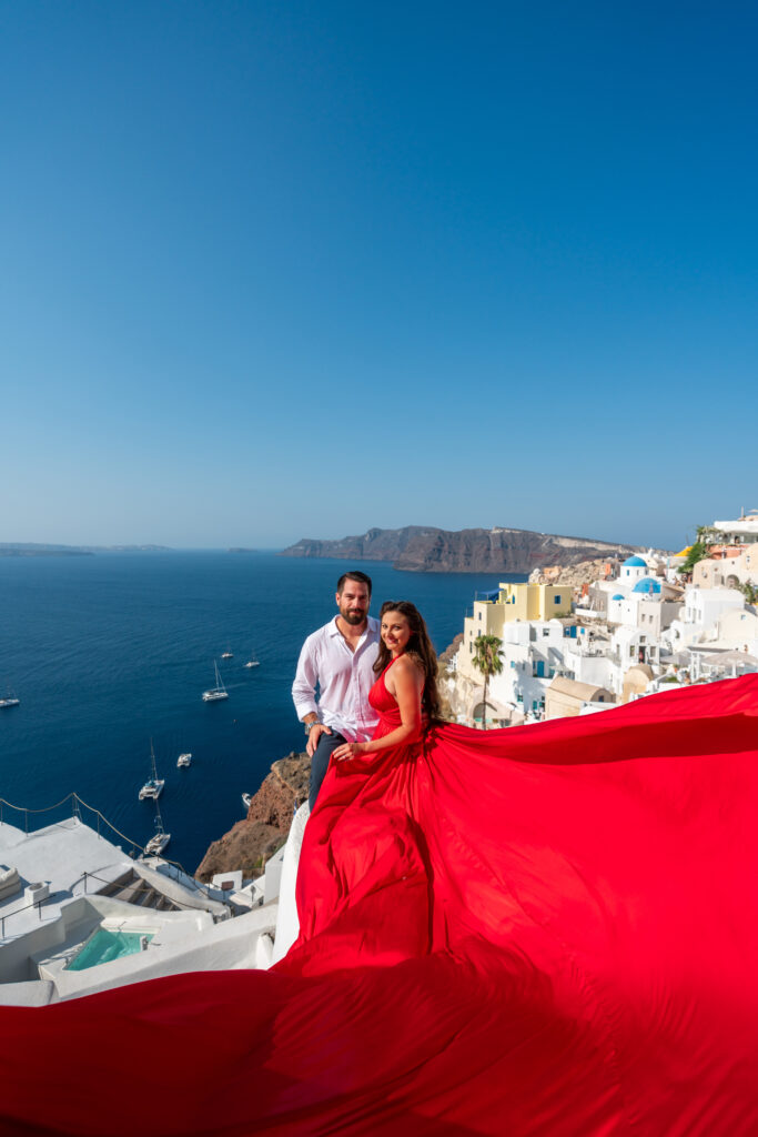 Woman wearing red flying dress poses with her husband in front of the Santorini caldera.