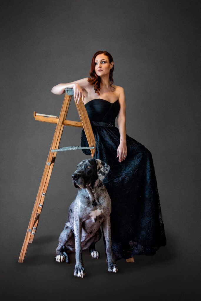 Vanity Fair style portrait of a woman in a black formal dress sitting on a wood ladder with her Great Dane at her feet.