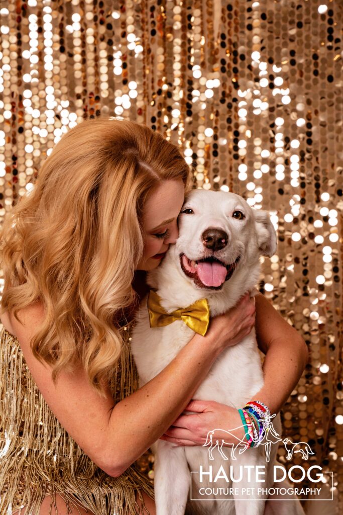 Woman with blonde hair hugs her white dog in front of a gold sequin backdrop.