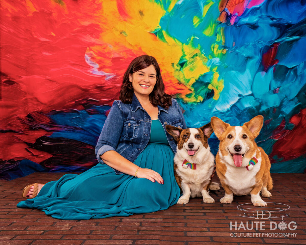 Woman sitting on a brick floor with two Corgis wearing rainbows on their collars in front of a rainbow painted backdrop.